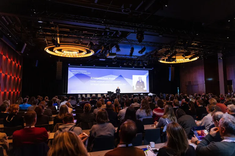 En sal med mennesker og en scene med lerret hvor presentatøren står